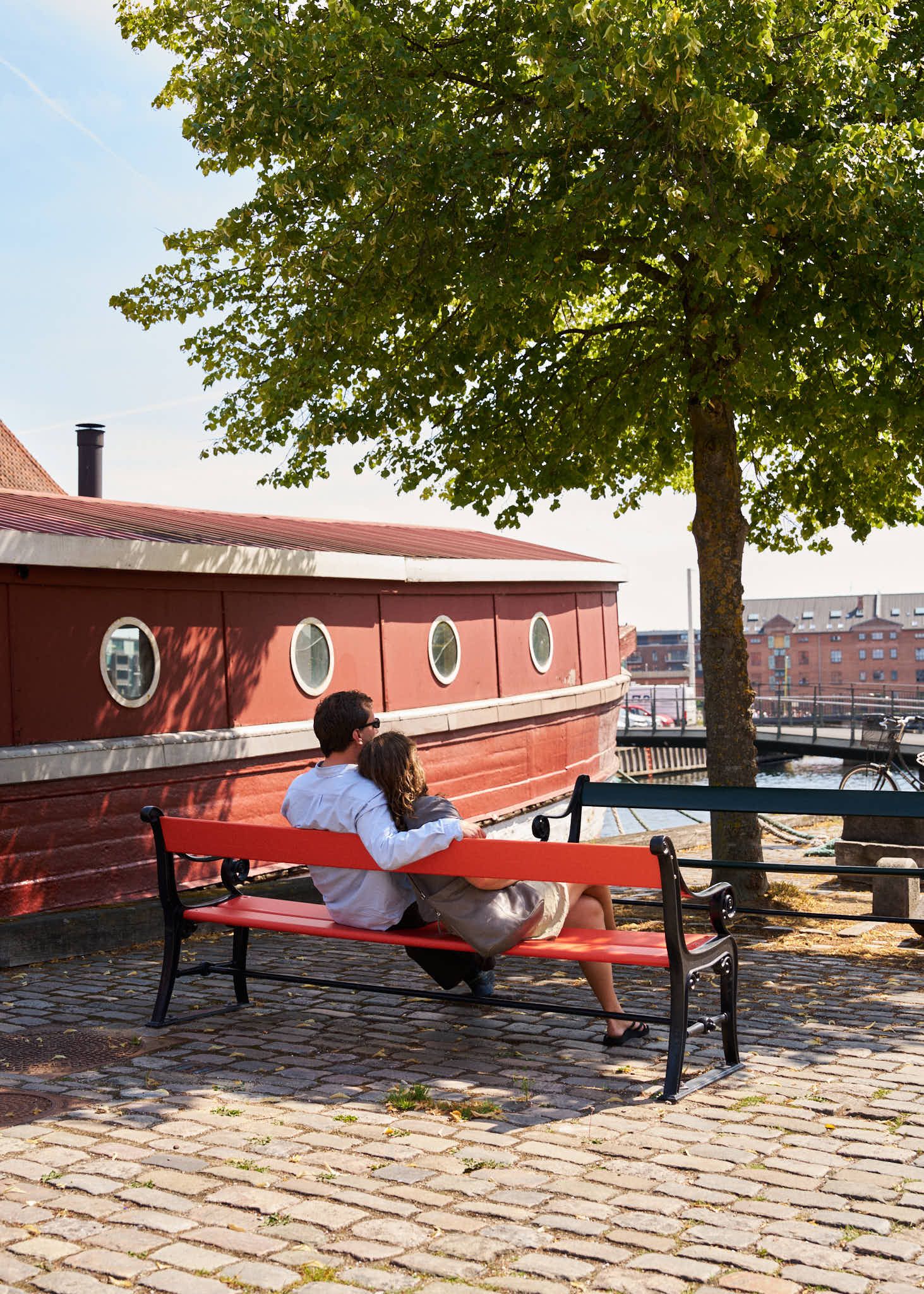 Two people on a red bench by a red houseboat on the Copenhagen harbour front