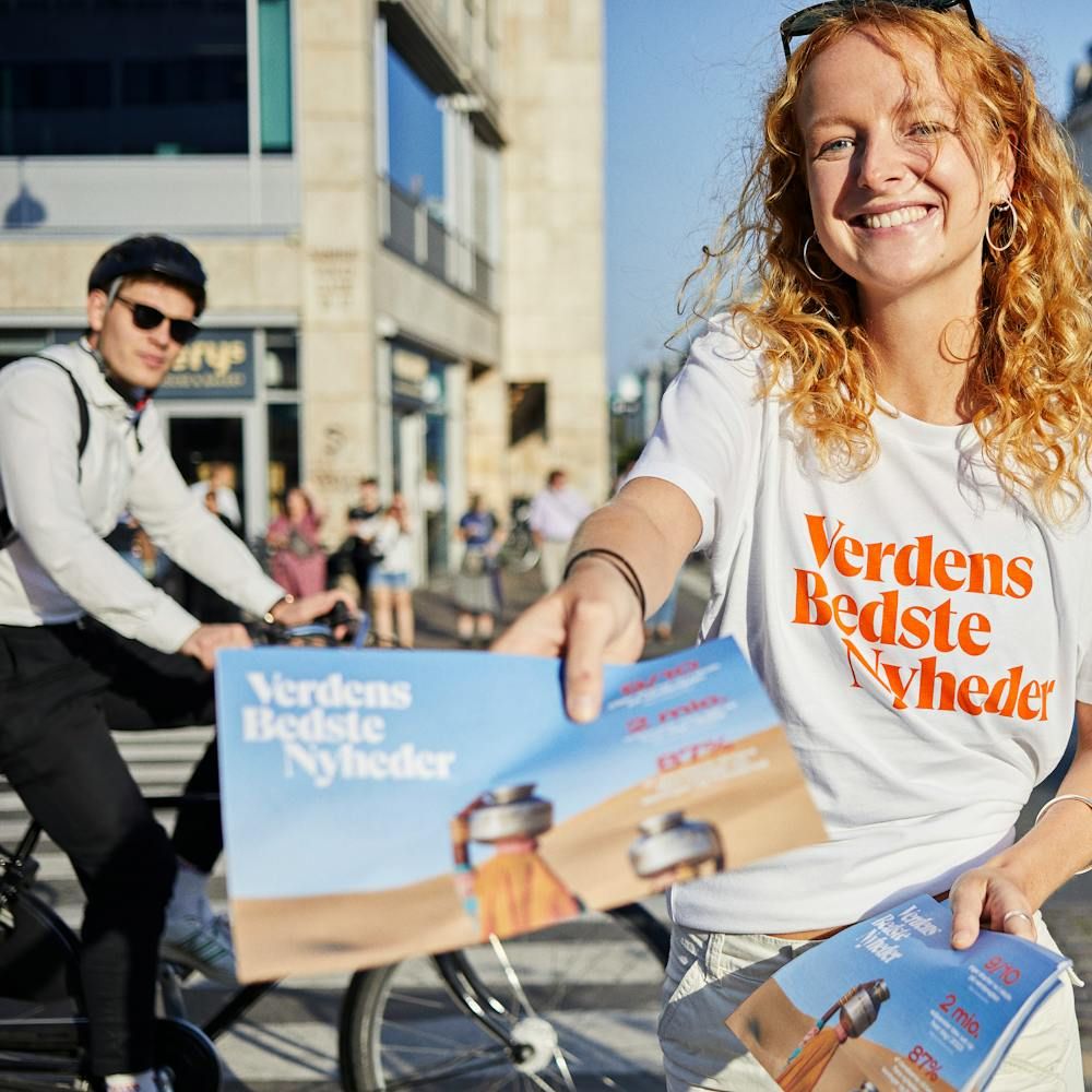 A smiling volunteer in a Verdens Bedste Nyheder T-shirt handing out newspapers to cyclists